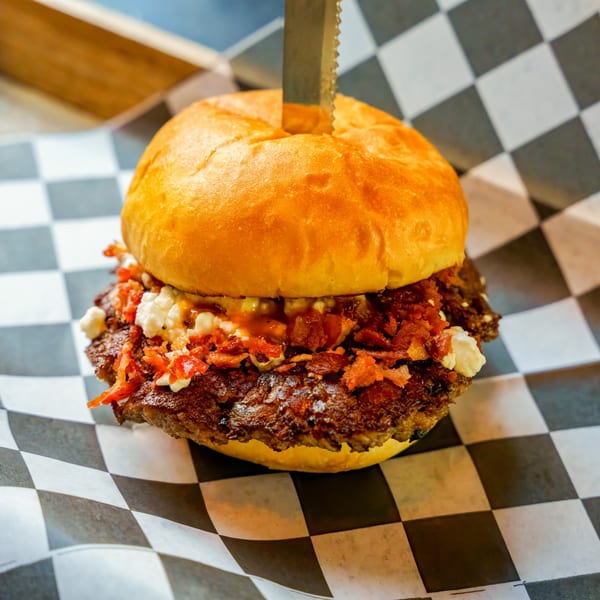 Lindy's on 4th burger with bacon, feta, and sauce, skewered with a knife on checkered paper.