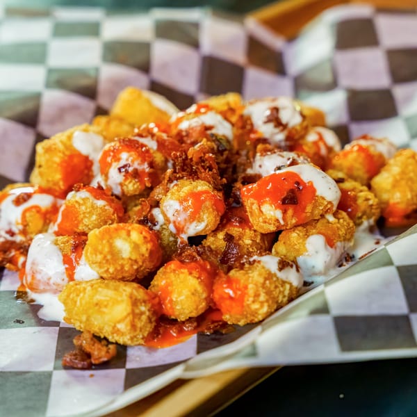 Golden tater tots topped with white sauce, red hot sauce, and bacon bits in a checkered paper-lined basket.