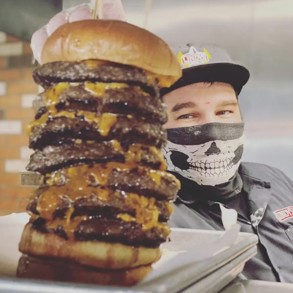 A Lindy's on 4th employee in a skull mask proudly presents a towering, multi-patty cheeseburger.
