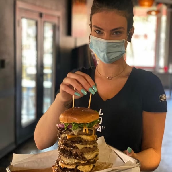 A masked Lindy's on 4th server holds a massive, multi-patty burger on a tray.