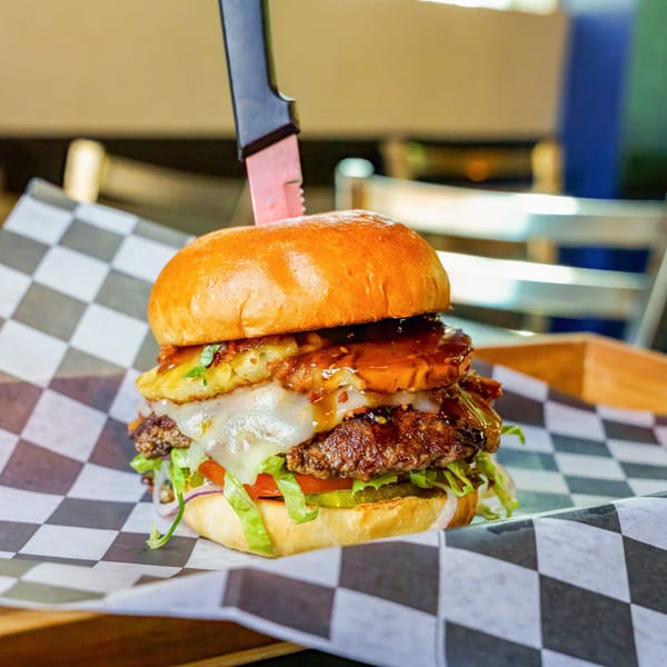 Lindy's on 4th burger with pineapple, cheese, and sauce, held by a knife on checkered paper.