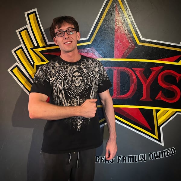 A man in glasses and a skull t-shirt stands smiling in front of a Lindy's on 4th logo.