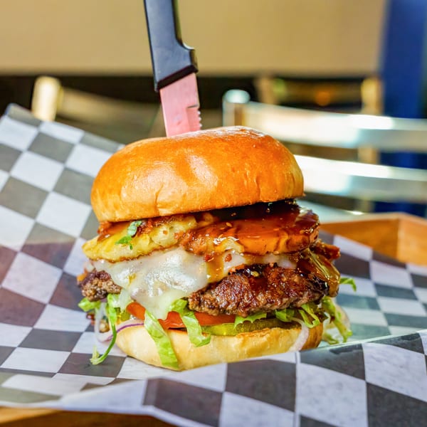 A Lindy's on 4th burger with melted cheese, pineapple, sauce, and vegetables, held by a knife.
