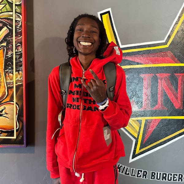 A smiling man in a red hoodie and backpack makes a rock-and-roll hand gesture in Lindy's on 4th.