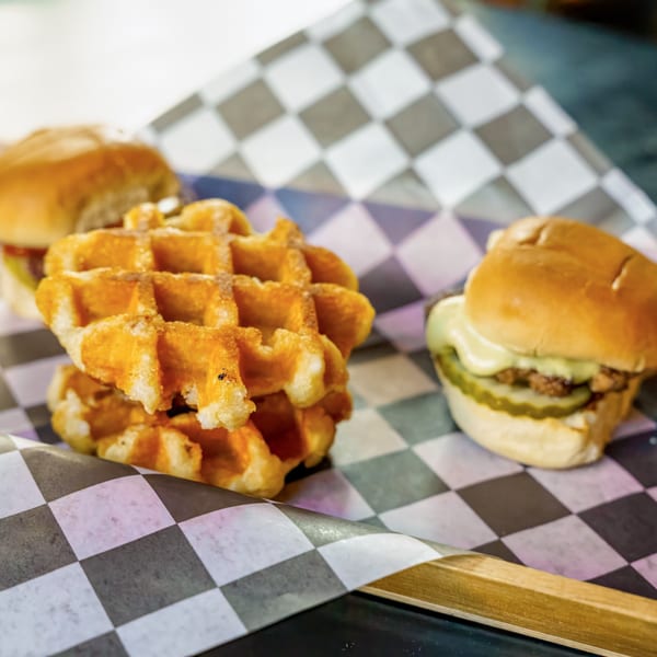 Two mini burgers with pickles and cheese, and a stack of waffles on checkered paper.