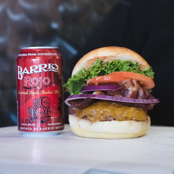 Lindy's on 4th burger with bacon, lettuce, tomato, and onion, next to a red Barrio Rojo beer can.