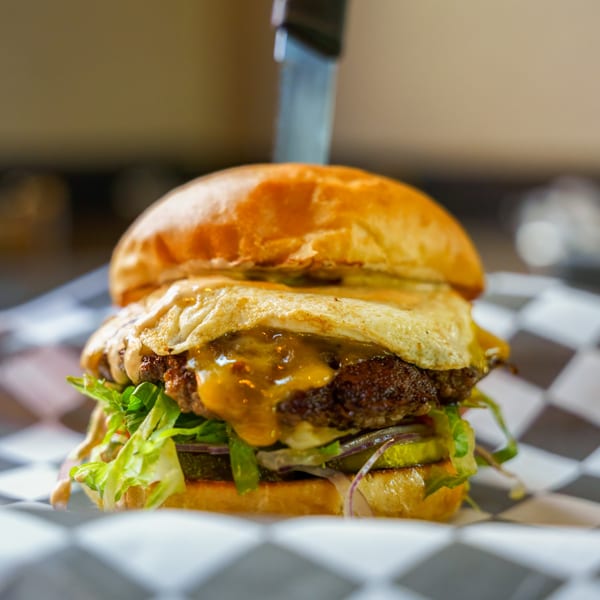 A Lindy's on 4th burger with a fried egg, melted cheese, lettuce, and pickles on a checkered wrapper.