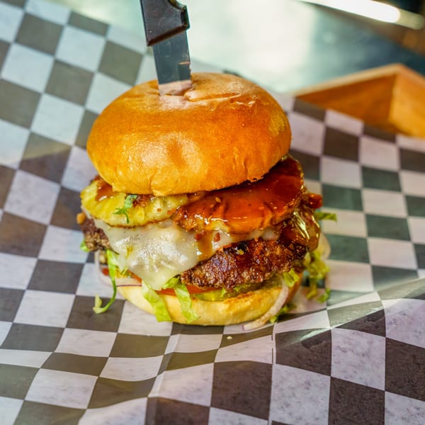 A Lindy's on 4th burger with pineapple, cheese, and sauce, held by a knife on checkered paper.