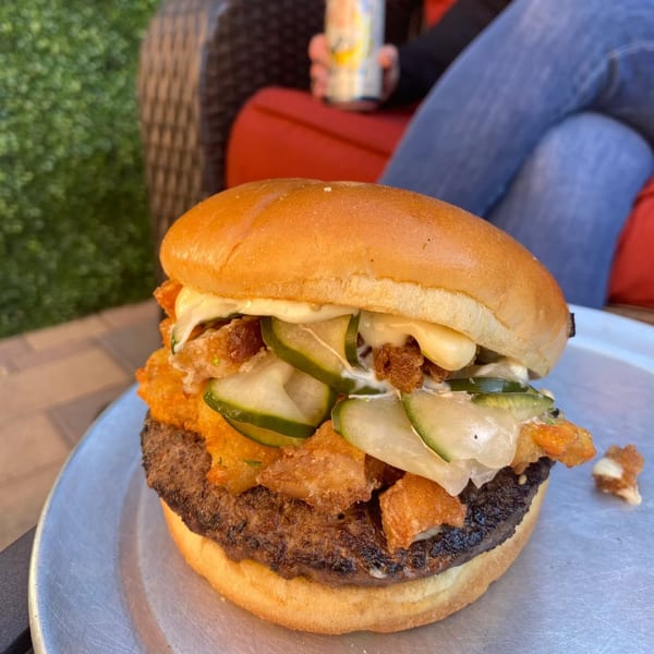A Lindy's on 4th burger with pickles, fried onions, and sauce on a metal plate outdoors.
