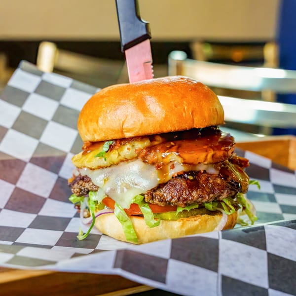 Lindy's on 4th burger with a knife, pineapple, melted cheese, and sauce on checkered paper.